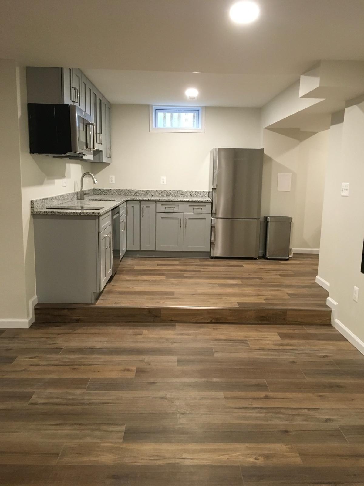 Basement kitchenette on a raised platform with gray cabinets and a stainless refrigerator