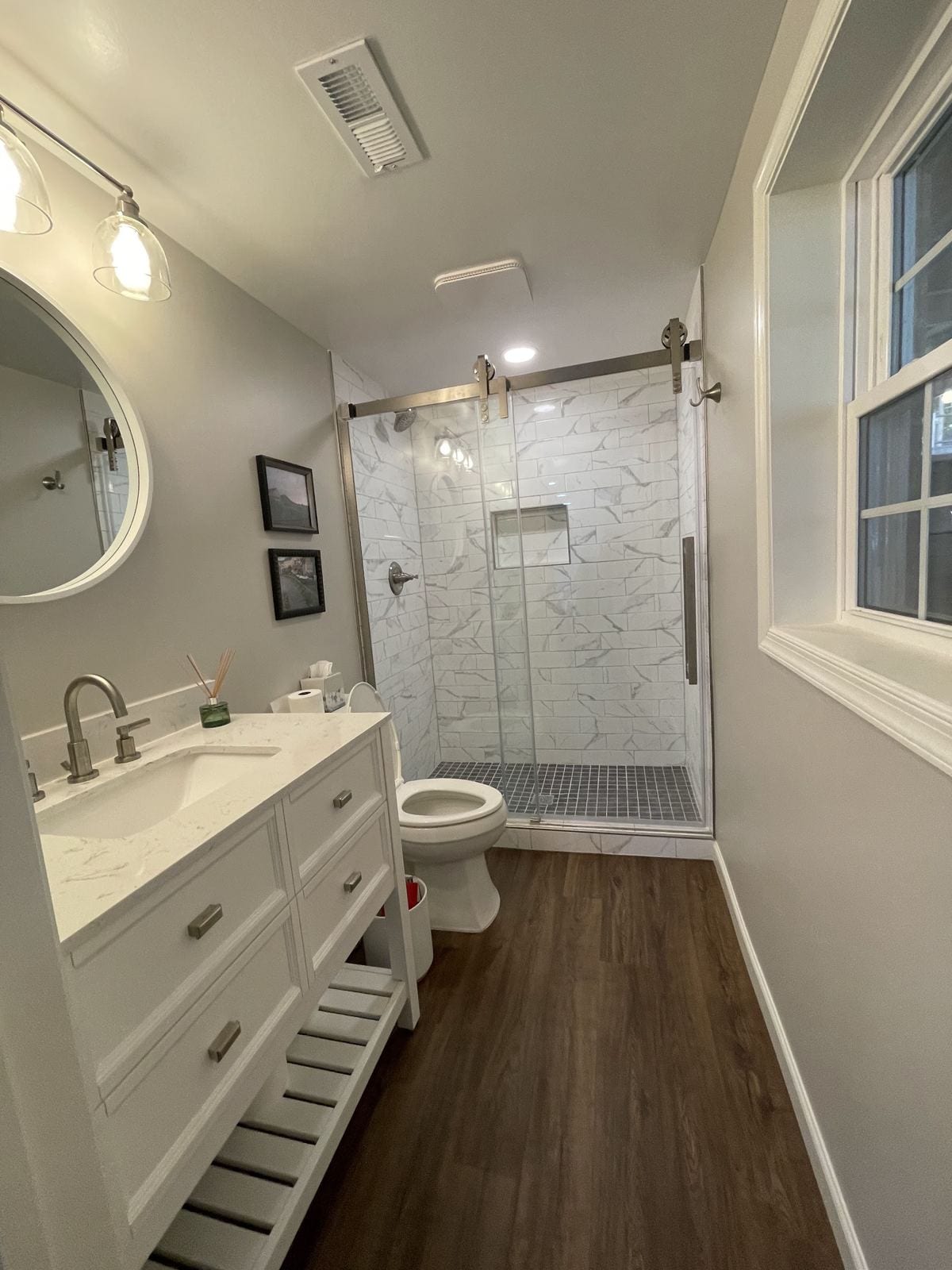 New basement bathroom with a white vanity, round mirror and marble-look tile shower